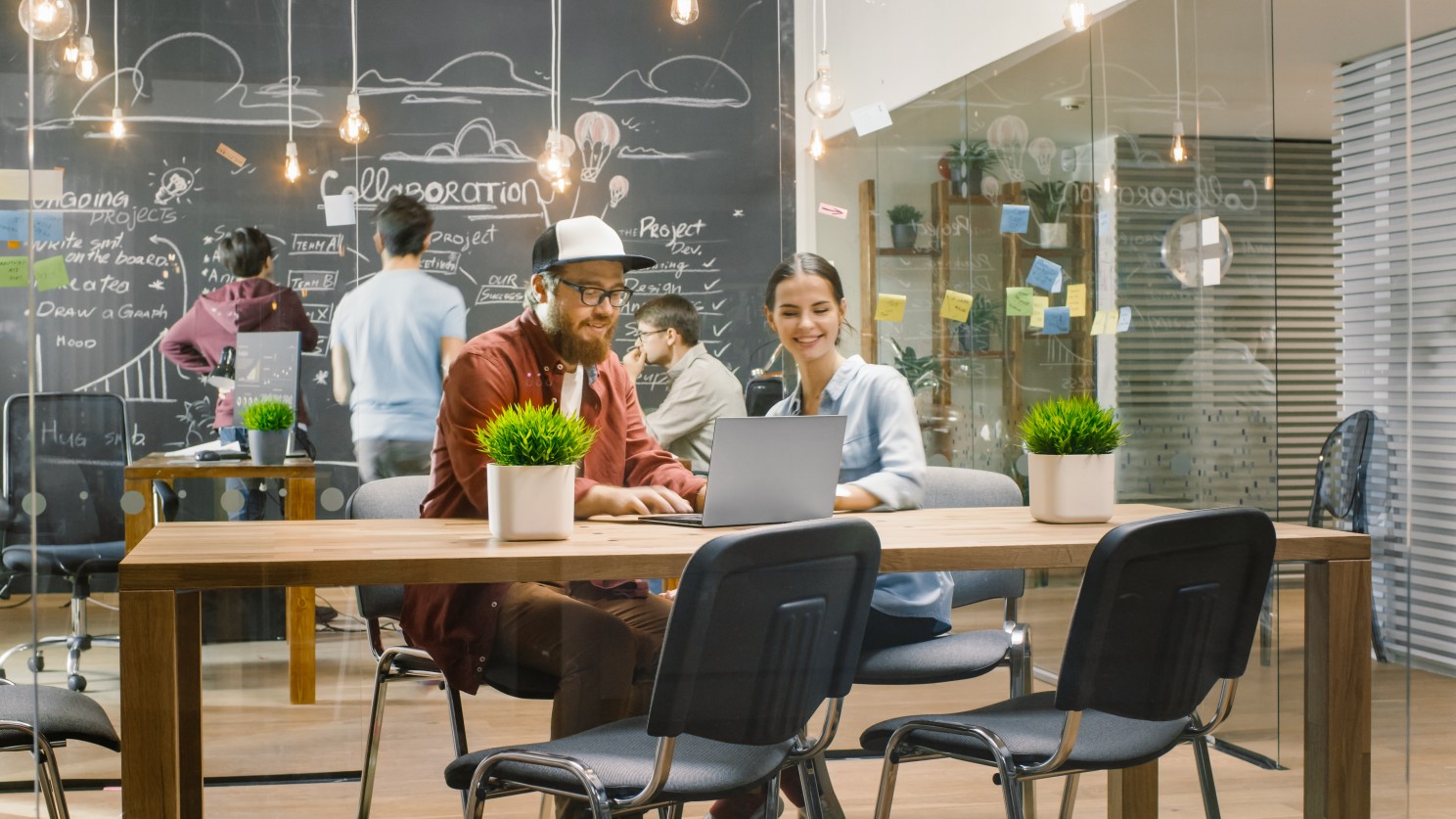 Two colleagues at a laptop in a loft office, with a chalkboard wall labeled Collaboration behind them