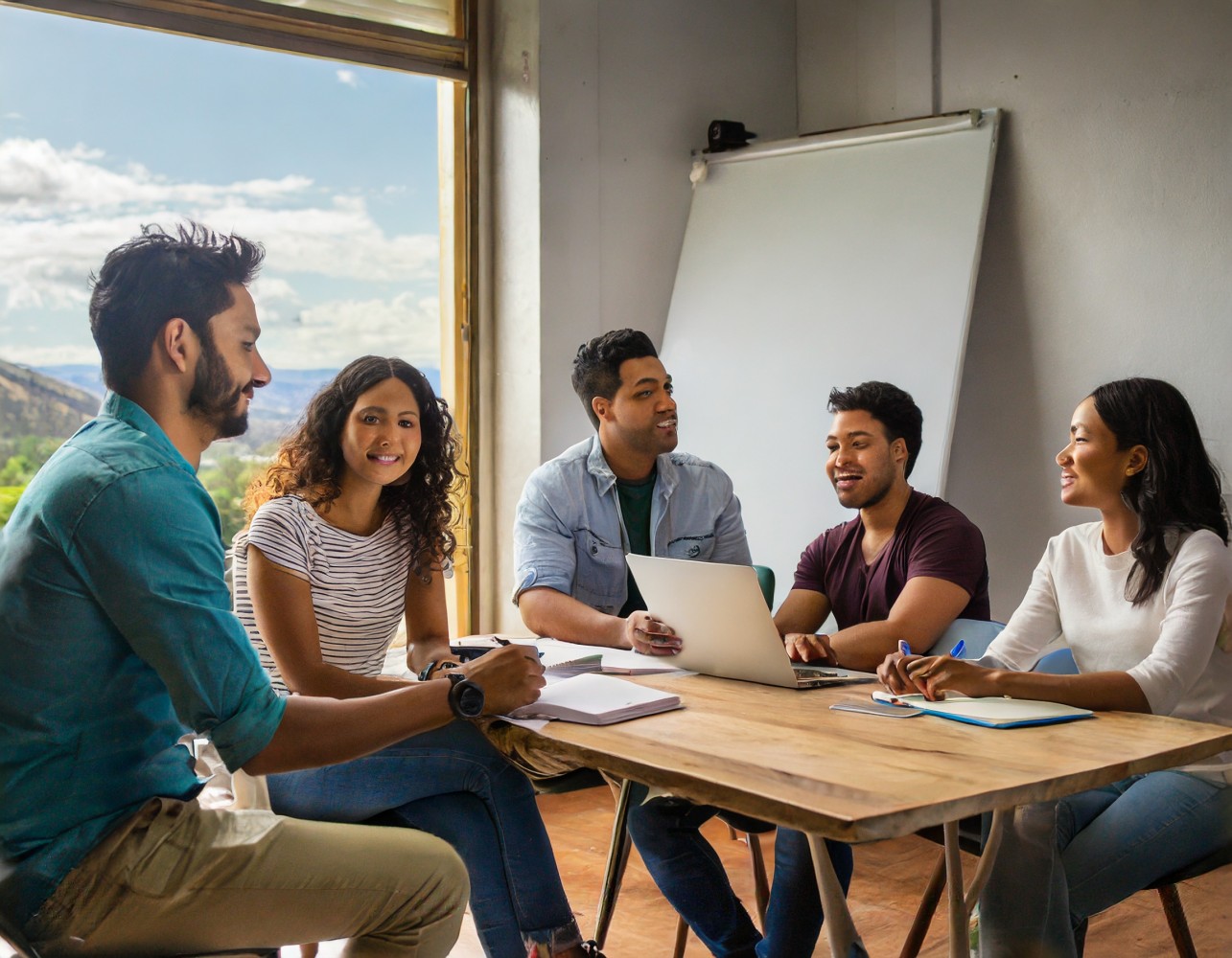 Five colleagues sharing knowledge around a wooden table with a laptop and notebooks beside a window