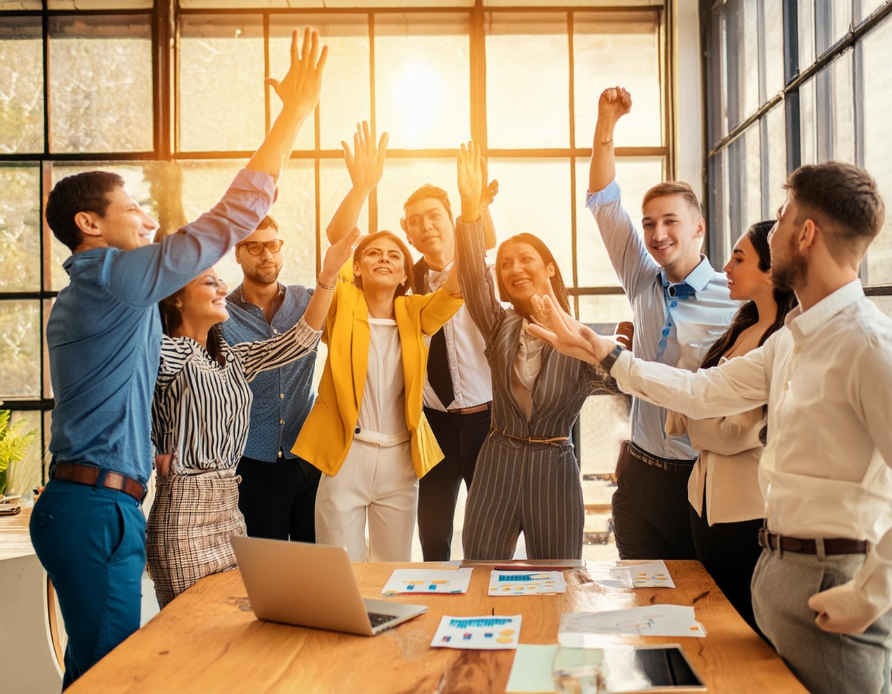 Team of coworkers raising their hands in a high-five celebration around a conference table in a sunlit office